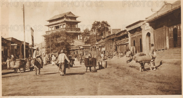 Street view in Beijing, page, photomechanical print, photographer: Donald Mennie, Beijing, cliché maker: anonymous, c. 1915 - in or before 1920, paper, rotogravure, height 160 mm x width 290 mm, street, A wide sepia street scene with a multi-tiered pagoda-style building rising behind rows of single-story shops and trees, people walking and standing along the street, several figures wearing loose garments and hats, a man riding a donkey and a person pushing a two-wheeled cart loaded with round baskets, a man carrying a yoke with buckets, wooden carts and bundles placed on small carts, a row of ornate wooden doors and arched entrances along the right side, simple poles and poles with cables, uneven dirt road surface and scattered shadows across the foreground.