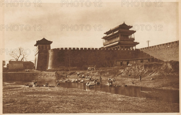 Unknown persons near a body of water along a city wall, page, photomechanical print, photographer: Donald Mennie, Beijing, cliché maker: anonymous, c. 1915 - in or before 1920, paper, rotogravure, height 179 mm x width 278 mm, historical persons, landscapes with waters, waterscapes, seascapes (in the temperate zone), city-walls, A sepia-toned scene shows a large stone wall with battlements and two multi-tiered pavilion structures rising above it, each with curved eaves. In front of the wall a sloped embankment leads down to a shallow river where several people are standing and crouching at the water's edge, some with bundles and some near small boats or rafts. Livestock and pack animals are present on the bank and near the water. On the left side a tree stands beside a smaller building adjacent to a tower incorporated into the wall. The foreground is an expanse of textured, bare ground stretching toward the river.