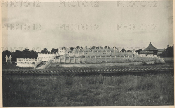 Altar of Heaven in Beijing, page, photomechanical print, photographer: Donald Mennie, Beijing, cliché maker: anonymous, c. 1915 - in or before 1920, paper, rotogravure, height 175 mm x width 280 mm, temple, shrine, non-Christian religions, Temple of Heaven, A wide black-and-white photograph of an ornate multi-tiered circular stone terrace with carved balustrades and staircases, set behind a low tiled wall and in front of a grassy foreground. The terrace features repeating decorative posts and relief panels along its tiers, with a wide central stairway leading upward. In the background there are dense dark-tree silhouettes and a pavilion-like building with a steep conical roof visible to one side. The sky above is pale and mostly empty.