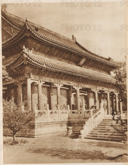 Summer Palace in Beijing, page, photomechanical print, photographer: Donald Mennie, Beijing, cliché maker: anonymous, c. 1915 - in or before 1920, paper, rotogravure, height 260 mm x width 204 mm, palace, Summer Palace, A sepia-toned photograph of a traditional multi-tiered building with ornate upturned eaves and tiled roofs, showing detailed carved brackets and decorative roof ridges. The facade features a row of tall columns supporting the overhanging eaves and intricately patterned window or door panels set between the columns. A stone balustrade runs along a raised platform in front of the building, interrupted by a wide flight of stone steps leading up to the entrance. Trees with sparse foliage frame the scene at the left and right edges, and shadowed ground stretches in the foreground.