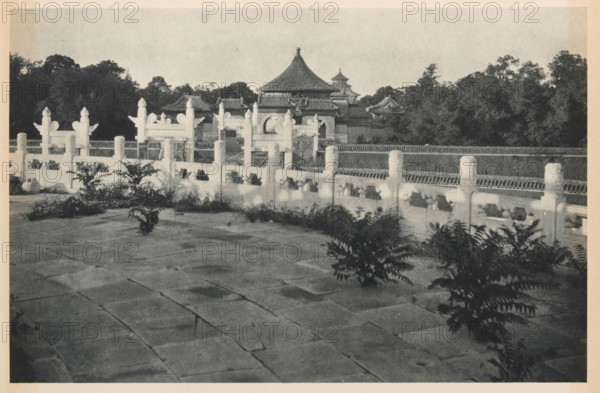 Altar of Heaven in Beijing, page, photomechanical print, photographer: Donald Mennie, Beijing, cliché maker: anonymous, c. 1915 - in or before 1920, paper, rotogravure, height 186 mm x width 278 mm, temple, shrine, traditional Chinese religions, Temple of Heaven, A wide stone courtyard with large rectangular paving stones fills the foreground, dotted with clusters of leafy plants. A low decorative balustrade with carved posts and openings runs across the middle ground, showing repeated ornamental pillars and rail sections. Behind the balustrade a series of traditional tiled-roof buildings rise, featuring curved eaves and a prominent multi-tiered roof with a pointed top. Dense trees and foliage form a dark backdrop behind the buildings under a pale sky. The image is monochrome with varied tones from light stone to dark vegetation.