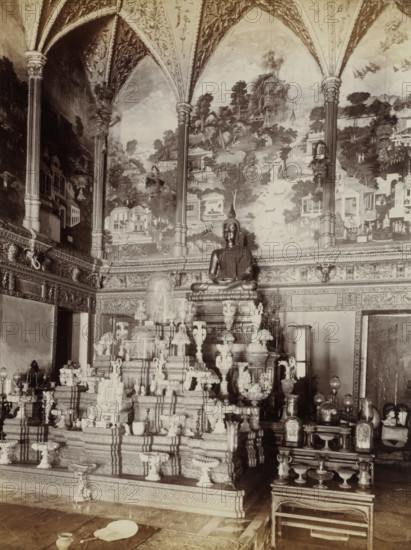 Interior of the Wat Pra Keo Temple (Temple of the Emerald Buddha) in Bangkok, album page, photograph, Part of Album with 50 photos of a trip through Southeast Asia., check stamp: 'GR Lambert Singapore', photographer: GR Lambert & Co., Bangkok, c. 1870 - c. 1900, paper, albumen print, height 273 mm x width 210 mm, temple, shrine, Hinduism, Buddhism, Jainism, A large seated statue on a raised ornate platform sits centrally beneath a vaulted ceiling with decorative arches and painted mural panels depicting trees, buildings, and landscape scenes, flanked by tall carved columns; the platform and surrounding area are filled with multiple intricately carved pedestals, vases, urns, and decorative objects arranged in tiers, and smaller altarpieces and covered displays occupy the foreground and sides, with patterned wall friezes and ornate molding running along the room, and a low mat and small vessels visible on the floor in front.