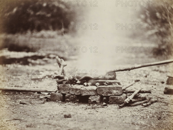 Cremation of the body of a boy in Siam, album page, photograph, Part of Album with 50 photos of a trip through Southeast Asia., negative number: '80', check stamp: 'GR Lambert & Co Singapore', photographer: GR Lambert & Co., Thailand, c. 1870 - c. 1900, paper, albumen print, height 209 mm x width 275 mm, body-burning, cremation, A sepia-toned photograph showing a rocky ground with a low stone platform in the center foreground, on top of which a human figure lies stretched out with legs raised and crossed at the ankles and arms extended alongside the body, a plume of pale smoke or steam rising vertically from the middle of the platform and obscuring the torso, a long wooden pole resting near the head and extending toward the right edge of the image, scattered logs and sticks around the platform, and a blurred natural background of foliage and uneven terrain.