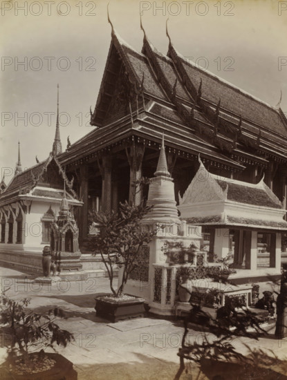 Main building of the temple complex of Wat Pra Keo (Temple of the Emerald Buddha) in Bangkok, album page, photograph, Part of Album with 50 photos of a trip through Southeast Asia., check stamp: 'GR Lambert Singapore', photographer: GR Lambert & Co., Bangkok, c. 1870 - c. 1900, paper, albumen print, height 274 mm x width 209 mm, temple, shrine, Hinduism, Buddhism, Jainism, A large ornate temple complex with steep multi-tiered gabled roofs decorated with upward-curving finials, flanked by smaller roofed structures and spires; a covered colonnade with carved columns and a raised platform; decorative stone and stucco elements including a stupa-like monument, an ornate shrine housing a small statue, and sculpted guardian figures; potted plants and small trees in square planters placed on a paved courtyard; shadowed foreground foliage and stone paving leading toward the buildings.