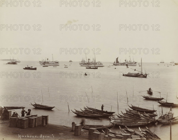 Harbour, S.pore, Ships and rowboats in the harbour of Singapore, album page, photograph, Part of Album with 50 photos of a trip through Southeast Asia., check stamp: 'GR Lambert Singapore', negative number: '6', photographer: GR Lambert & Co., Singapore, c. 1870 - c. 1900, paper, albumen print, height 210 mm x width 268 mm, A wide view of a harbor scene showing calm water with numerous boats and ships at varying distances, small wooden rowboats and narrow longboats clustered near the foreground alongside a stone quay where crates and bollards are visible, several people standing or sitting on the quay and on some boats, a few figures rowing in small boats on the water, a larger sailboat with a dark sail and several steam or motor ships farther out, slender poles rising from the water among the boats, and a pale sky above the horizon.