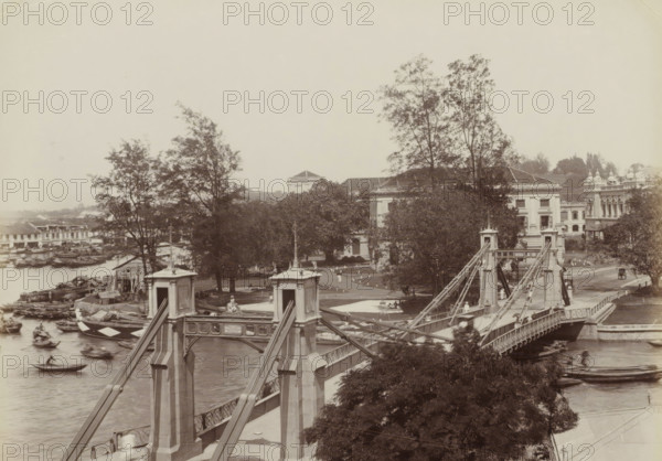 Cavenach Bridge, S'pore, Cavenach Bridge in Singapore, album page, photograph, Part of Album with 50 photos of a trip through Southeast Asia., check stamp: 'GR Lambert Singapore', negative number: '10', photographer: attributed to GR Lambert & Co., Singapore, c. 1870 - c. 1900, GR Lambert & Co., photographer, Attributed to, paper, albumen print, height 209 mm x width 274 mm, bridge in city across river, canal, A sepia-toned scene of a suspension bridge crossing a river, with ornate stone towers and metal cables, several boats moving in the water below, and a riverside area with small wooden structures and stacked timber. On the far bank there are trees with dense foliage partially obscuring large classical-style buildings with columns and tiled roofs. People appear as small figures on the bridge and along pathways near the buildings, and a carriage is visible on a road beside the riverside structures. The sky is pale and empty, and foreground foliage frames the lower part of the image.
