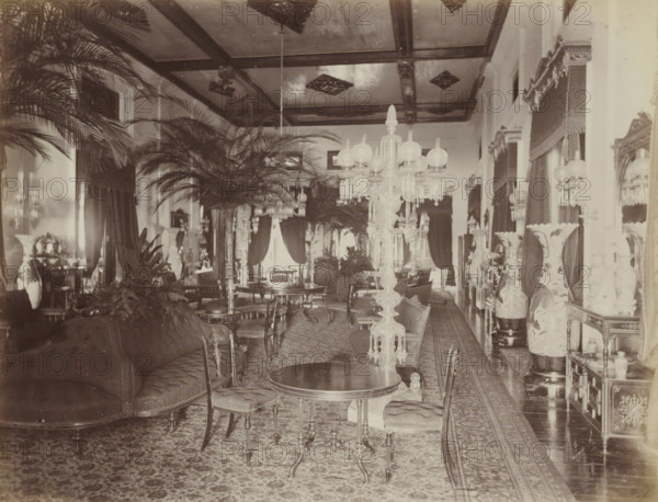 Salon in the palace of the Sultan of Johor, album page, photograph, Part of Album with 50 photos of a trip through Southeast Asia., check stamp: 'GR Lambert Singapore', photographer: GR Lambert & Co., Malaysia, c. 1870 - c. 1900, paper, albumen print, height 211 mm x width 275 mm, drawing-room, 'salon', A long ornate room filled with upholstered sofas and wooden chairs around circular and oval tables, tall decorative floor lamps with glass shades and hanging crystal prisms, large potted palm plants and other foliage arranged along the center and sides, patterned carpets covering the floor, tall vases with painted designs on stands along one side, heavy draped curtains framing tall windows, large mirrors and framed openings on the walls, a coffered ceiling with decorative panels, and various small decorative objects and furniture pieces placed throughout the space.
