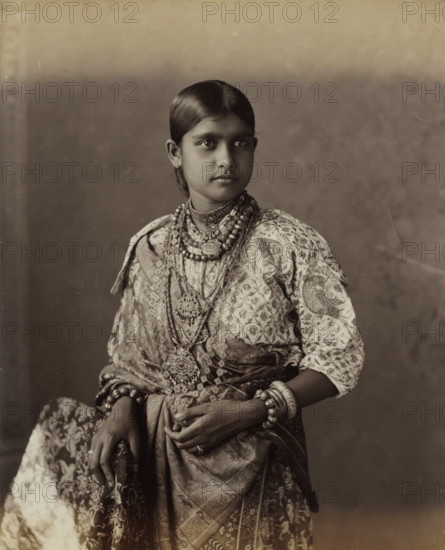 Kandian Lady, Portrait of a young woman from Kandy in Ceylon, album page, photograph, Part of Album with 50 photographs of a trip through Southeast Asia., photographer: Charles T. Scowen, Sri Lanka, c. 1875 - c. 1880, South Asia, paper, albumen print, height 278 mm x width 216 mm, adolescent, young woman, maiden, A young person with neatly parted dark hair and a calm expression is shown from the waist up wearing patterned clothing and multiple layered necklaces and pendants, bracelets and bangles on both wrists, and a patterned wrap draped across the body, with hands resting in front and a plain backdrop behind.
