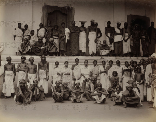 Men and women posing in front of a building, possibly in Ceylon, album page, photograph, Part of Album with 50 photos of a trip through Southeast Asia., stamp: 'Fabrikmarke', photographer: attributed to Charles T. Scowen & Co., Sri Lanka, c. 1875 - c. 1880, South Asia, Attributed to, paper, albumen print, height 218 mm x width 277 mm, clothes, costume, A large group of people arranged in tiers in front of a building, many standing on a raised platform and others seated or squatting on the ground, wearing traditional wrapped garments and headwraps, with varied bare torsos and some shawls draped over shoulders; the background shows a plain wall with doorways and steps, and the faces show a range of neutral expressions, while some individuals rest arms on knees or cross their arms, and a few hold simple objects or cloths.