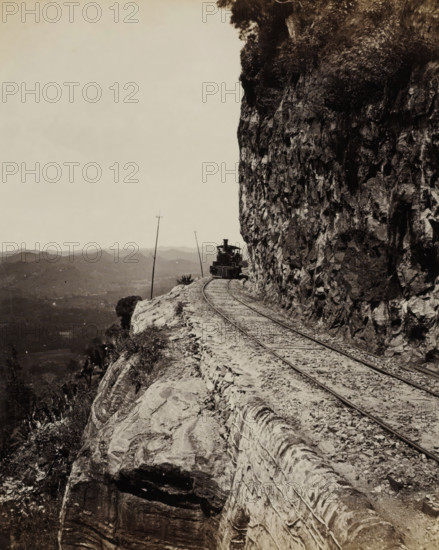 Train runs along a precipice at Sensation Rock in Ceylon, Sensation Rock. Colombo and Kandy Line., album page, photograph, Part of Album with 50 photos of a trip through Southeast Asia., photographer: Charles T. Scowen & Co., Sri Lanka, c. 1876 - c. 1885, South Asia, paper, albumen print, height 277 mm x width 217 mm, railway, train, mountains, A narrow railway track runs along a rocky cliff edge with a steam locomotive visible ahead on the track, partially framed by the cliff face on the right; the cliff shows jagged rock textures and vegetation at the top, while the left side opens to a distant hilly landscape under a pale sky and a couple of leaning telegraph poles rise near the track.