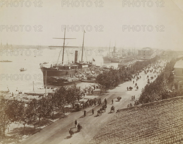 View of the Bund waterfront in Shanghai, album page, photograph, Part of Album with 50 photographs of a trip through Southeast Asia., Shanghai, c. 1860 - c. 1900, photographer, paper, albumen print, height 212 mm x width 270 mm, harbour, A wide waterfront scene shows a busy quay lined with large moored ships with tall masts and smokestacks, smaller boats and barges alongside, and piled cargo on the docks; a broad tree-lined street runs parallel to the water filled with many pedestrians, horse-drawn carriages, and figures moving in different directions; buildings and warehouses sit near the docks, and the far water is dotted with more ships and small boats under a pale sky.
