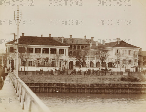 Exterior of the Astor House Hotel in Shanghai, album page, photograph, Part of Album with 50 photos of a trip through Southeast Asia., Shanghai, in or after 1859 - c. 1900, photographer, paper, albumen print, height 204 mm x width 274 mm, hotel, hostelry, inn, A sepia-toned photograph shows a large two-story building with a tiled roof and multiple chimneys, featuring an open upper veranda with columns and an arched central entrance flanked by windows and smaller arches; leafless trees stand in front of the building and a fenced lawn separates it from a body of water in the foreground, with a wooden bridge and railing extending along the left side of the image; several horse-drawn carriages and groups of people are visible near the fence and entrance, and a tall utility pole with crossarms stands near the left front of the building.