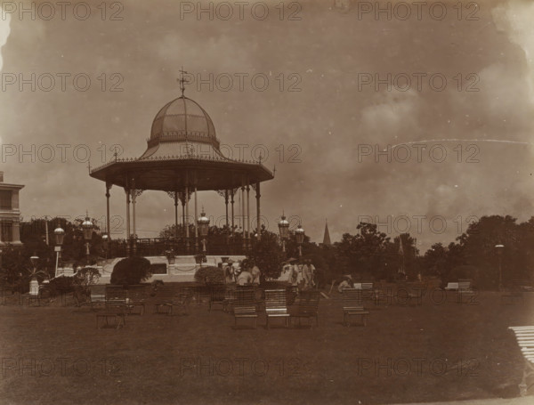 Public gardens in Shanghai, album page, photograph, Part of Album with 50 photos of a trip through Southeast Asia., Shanghai, c. 1860 - c. 1900, photographer, paper, albumen print, height 195 mm x width 252 mm, public gardens, park, A large ornate bandstand with a domed roof and decorative spire stands on a raised platform supported by slender columns, surrounded by lamp posts with embellished lanterns; in front of it are rows of empty wooden slatted chairs on a grassy lawn, low hedges and shrubs, and a few people gathered near the steps; trees and a peaked steeple are visible behind the structure, and a multi-story building appears at the far left.