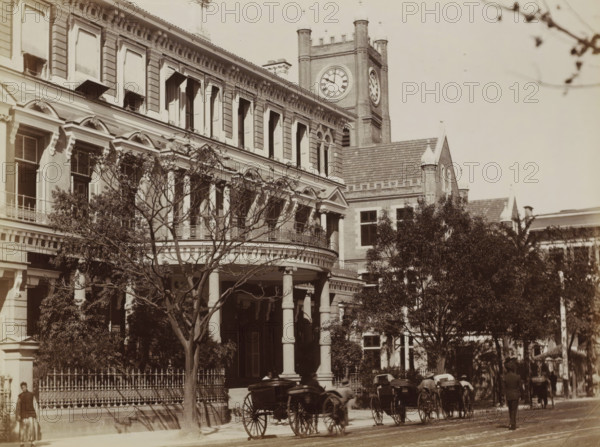 Exterior of the Hong Kong-Shanghai Bank on the Bund in Shanghai, album page, photograph, Part of Album with 50 photographs of a trip through Southeast Asia., Shanghai, c. 1860 - c. 1900, photographer, paper, albumen print, height 201 mm x width 270 mm, exterior, representation of a building, A sepia-toned street scene showing a large multi-story building with ornate classical architecture, arched windows with shutters, decorative cornices, and a curved balcony supported by columns, trees with bare and leafy branches partially obscuring the facade, a tall clock tower rising behind the building with visible clock faces, a wrought-iron fence along the sidewalk, several horse-drawn carriages parked or moving on the street, and a few pedestrians walking along the pavement.