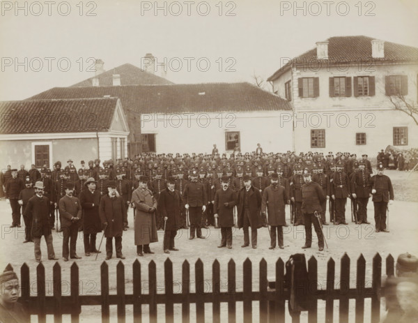 Group portrait of Western policemen of the Shanghai International Concession, album page, photograph, Part of Album with 50 photos of a trip through Southeast Asia., Shanghai, c. 1880 - c. 1910, photographer, paper, albumen print, height 212 mm x width 271 mm, policeman, constable, A large group of men arranged in rows stands in front of a cluster of light-colored buildings with tiled roofs and shuttered windows; many of the men wear dark uniforms and peaked or bowler hats, some hold canes or swords, and a few wear long overcoats and facial hair; behind them a few taller figures stand near windows and doorways, and a wooden picket fence runs across the foreground.