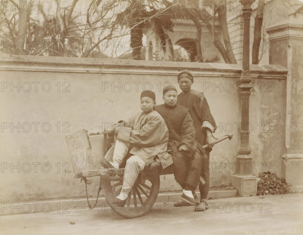 Three Chinese people in Shanghai use a wheelbarrow as a means of transport, album page, photograph, Part of Album with 50 photos of a trip through Southeast Asia., address: '42 Nanking Road', photographer: Sze Yuen Ming & Co, Shanghai, c. 1860 - c. 1900, paper, albumen print, height 212 mm x width 270 mm, wheelbarrow, Three people are positioned around a wooden cart with a large spoked wheel in front of a stone wall; two sit on the cart while one stands behind holding the cart's handle. The seated individuals wear long, loose garments and round caps, and the standing person wears a long dark robe with a wrapped head covering and sandals. The cart shows visible wood grain and rope attachments, and a circular pattern is faintly visible on the cart's side. Behind them, a wall topped with a ledge runs horizontally across the scene, with leafless tree branches and parts of a building visible above the wall. A tall ornate lamppost or column stands to the right against the wall, and a small p...
