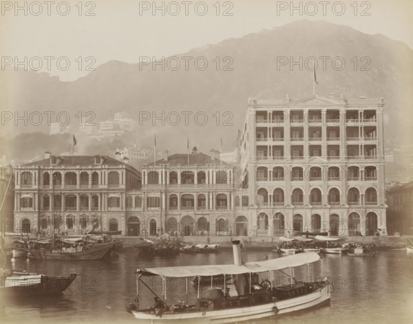 View of the Hong Kong Hotel in Hong Kong, album page, photograph, Part of Album with 50 photos of a trip through Southeast Asia., Hong Kong, c. 1860 - c. 1900, photographer, paper, albumen print, height 223 mm x width 284 mm, hotel, hostelry, inn, A waterfront scene showing a row of ornate multi-story buildings with arched ground-floor openings, columns, balconies, and decorative facades, with a taller building on the right and lower connected buildings to the left; small boats and sampans are moored in the water in the foreground, one larger covered boat with a canopy and visible equipment is closest to the viewer; hills rise behind the buildings, with additional structures partially visible on the slopes and a cross-shaped structure near the hilltop; the overall image has a sepia tone and a hazy atmosphere.