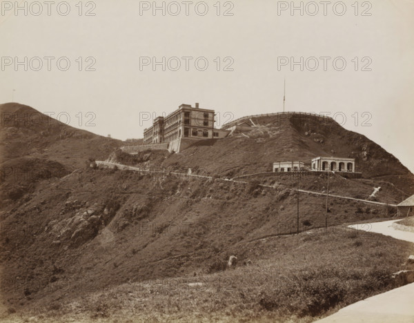 Buildings on the top of The Peak (Mount Austin) in Hong Kong, album page, photograph, Part of Album with 50 photos of a trip through Southeast Asia., Hong Kong, c. 1860 - c. 1900, photographer, paper, albumen print, height 213 mm x width 272 mm, railway, train, mountains, A sepia-toned hillside scene showing a large multi-story building with rows of windows and balconies positioned on a terraced slope, a smaller pavilion-like structure with open arches located lower on the hill, winding paths and retaining walls that trace the contours of the terrain, rocky outcrops and grassy vegetation covering the slopes, a tall thin pole or mast rising from the ridge near a low fence, and a pale sky occupying the upper part of the image.