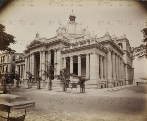 Exterior of the Hong Kong-Shanghai Bank in Hong Kong, album page, photograph, Part of Album with 50 photographs of a trip through Southeast Asia., Hong Kong, c. 1860 - c. 1900, photographer, paper, albumen print, height 233 mm x width 288 mm, exterior, representation of a building, A large neoclassical stone building with a central dome and a cupola rises above a colonnaded facade, showing tall fluted columns with ornate capitals supporting a heavy entablature and balustraded roofline, decorative pediments and sculptural urns along the roof, and an open arched entrance beneath the portico; young palm trees in metal guards line the sidewalk in front of the building, parts of adjacent classical buildings are visible at the sides, a paved street runs in front with a covered bench or cart and a small section of a chair in the foreground, and leafy tree branches frame the left and right edges of the scene.