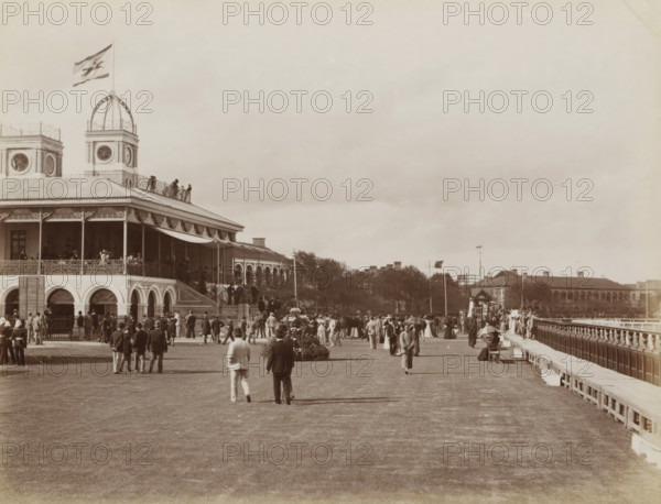 View of the horse racetrack and grandstand of Shanghai, album page, photograph, Part of Album with 50 photos of a trip through Southeast Asia., Shanghai, c. 1890 - c. 1900, photographer, paper, albumen print, height 215 mm x width 277 mm, horse-racing, race-track, A large two-story building with arched openings on the ground level and a covered upper veranda occupies the left side, topped by a tower with a round window and an open dome structure bearing a flag, with people visible on the veranda and along a railing on the roof; in front of the building a wide open lawn stretches toward the foreground where groups of men and women in period clothing are standing, walking, and gathering, some wearing hats and some holding umbrellas, with floral plantings and a raised pathway or promenade flanked by a balustrade on the right leading toward distant low buildings and trees under a broad cloudy sky.