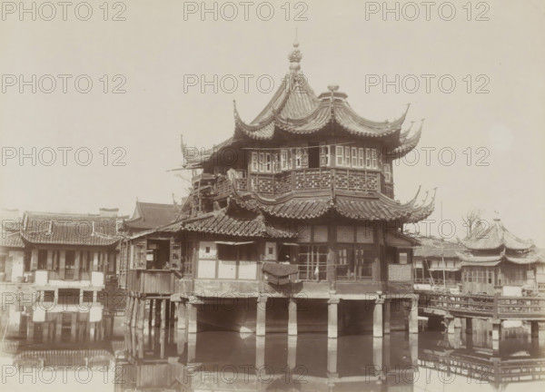Mandarin teahouse in Chinatown Shanghai, album page, photograph, Part of Album with 50 photos of a trip through Southeast Asia., Shanghai, c. 1860 - c. 1900, photographer, paper, albumen print, height 197 mm x width 268 mm, inn, coffee-house, public house, A multi-tiered wooden pavilion with sweeping curved roofs and ornate eaves stands on raised stone pillars above a body of water, showing carved lattice windows, tiled roof ridges, and decorative finials atop the roof; adjacent lower buildings with similar tiled roofs and open windows flank the pavilion, and a covered wooden walkway and small bridge connect parts of the complex, with reflections of the structures visible in the water below.