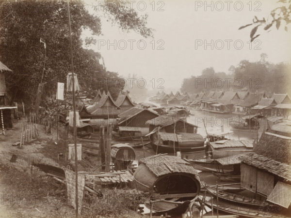 Boats at the quay of a waterway in Bangkok, album page, photograph, Part of Album with 50 photos of a trip through Southeast Asia., check stamp: 'GR Lambert & Co Singapore', photographer: GR Lambert & Co., Bangkok, c. 1870 - c. 1900, paper, albumen print, height 207 mm x width 274 mm, river bank, A riverside scene filled with numerous wooden boats and houseboats clustered along the water, many with thatched roofs and curved woven coverings, narrow walkways and wooden posts among them, a line of low houses with steep gabled roofs running along the far bank, dense trees and foliage rising behind the buildings, a dirt path and simple wooden structures at the near left edge, tall poles and hanging cloths or bundles visible near the shore, overhanging branches and leaves framing the top of the image, and a hazy sky above.