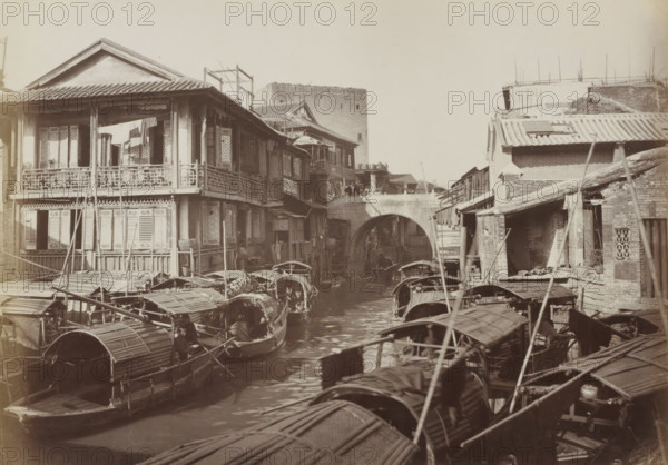 Boats and houses in Canton, album page, photograph, Part of Album with 50 photos of a trip through Southeast Asia., Canton (city), c. 1860 - c. 1900, photographer, paper, albumen print, height 205 mm x width 291 mm, harbour, A narrow waterway crowded with small covered wooden boats stretches toward a stone arched bridge, flanked on both sides by multilevel riverside buildings with tiled roofs and wooden balconies; the left bank shows a larger building with decorative railing and open windows with fabric hanging on the upper balcony, while the right bank has smaller brick and wooden structures with sloped roofs and exposed beams; boats with curved canopies and long poles fill the foreground and middle distance on the water, and a tall rectangular masonry tower rises behind the buildings in the background.