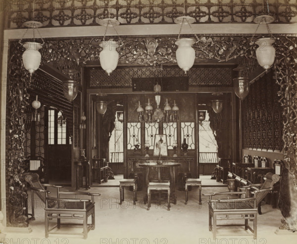 Interior of a Chinese club in Guangzhou, album page, photograph, Part of Album with 50 photos of a trip through Southeast Asia., Guangzhou (city), c. 1860 - c. 1900, photographer, paper, albumen print, height 228 mm x width 275 mm, club, meeting place, An interior room with ornate carved woodwork framing the entrance and decorative patterned screens along the back wall, suspended light fixtures with glass shades hanging from the ceiling, a central round table with stools around it and a chandelier above, pairs of wooden armchairs with open lattice seats positioned near the foreground, additional small tables and cabinets arranged along the sides, carved decorative panels and hanging lanterns visible throughout, and windows with patterned panes letting light into the space.
