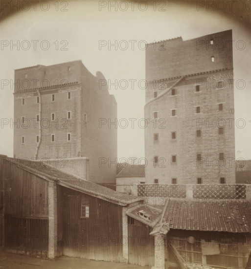 Buildings of pawnbrokers in Guangzhou, album page, photograph, Part of Album with 50 photos of a trip through Southeast Asia., Guangzhou (city), c. 1860 - c. 1900, photographer, paper, albumen print, height 258 mm x width 234 mm, pawnbroker, lombard, Two tall brick towers with small rectangular openings rise behind a cluster of lower wooden and tiled-roof buildings; the towers show patterned brickwork near their tops and a vertical drainpipe on one face, while the foreground roofs display rows of curved tiles, exposed rafters, and weathered wooden planks, and a lattice brick wall runs between parts of the buildings.