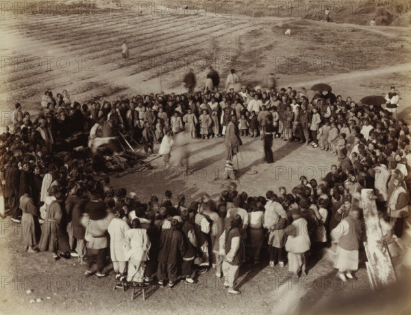 Chinese people in a circle at an outdoor board game, album page, photograph, Part of Album with 50 photos of a trip through Southeast Asia., title: 'Board game', number: '550', China, c. 1860 - c. 1900, photographer, paper, albumen print, height 214 mm x width 278 mm, sports, games and physical performances, A large crowd of people stands and sits outdoors arranged in a rough circle around a central open area where a few figures stand and one person is seated on the ground with nearby bowls and simple objects, many people wear layered garments and head coverings, some hold umbrellas, and several small children are visible among the adults; in the background there are terraced earth beds and a sloped grassy area with a few scattered individuals and a small dog, and the scene is captured in a sepia-toned photograph.