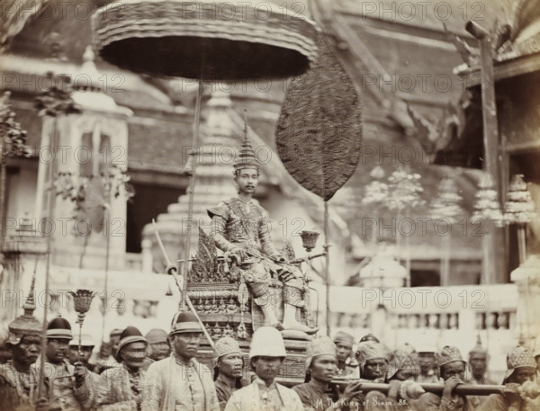 HM The King of Siam, Procession with Rama V, King of Siam, on a palanquin, album page, photograph, Part of Album with 50 photos of a trip through Southeast Asia., check stamp: 'GR Lambert & Co Singapore', negative number: '85', photographer: GR Lambert & Co., Thailand, c. 1870 - c. 1900, paper, albumen print, height 210 mm x width 274 mm, Person, Chulalongkorn the Great, ceremonial procession, parade, pageant, installation of a ruler (variant), A sepia-toned scene shows a central seated figure wearing ornate patterned clothing and a tall pointed headpiece, positioned on an elaborately decorated palanquin. Above the seated figure is a large round parasol and a tall textured fan held on poles. Surrounding the palanquin are numerous men dressed in traditional attire and hats, some carrying poles or ceremonial objects, their faces visible in the foreground. Architectural elements such as stepped structures and ornate building facades are visible in the background, with decorative details a...