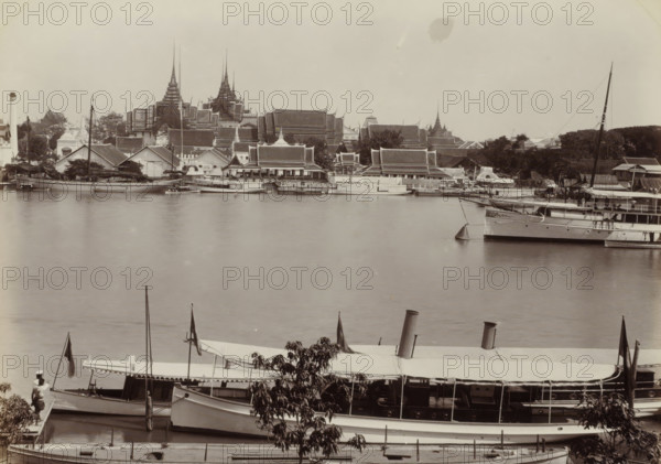 Menam River in Bangkok with buildings and boats, seen from the Royal Dock Yard, Menam River from the Royal Dock Yard, B, ok, album page, photograph, Part of Album with 50 photos of a trip through Southeast Asia., negative number: '45', check stamp: 'GR Lambert Singapore', photographer: GR Lambert & Co., Bangkok, c. 1870 - c. 1900, paper, albumen print, height 210 mm x width 275 mm, pier, quay, wharf, A wide riverside scene showing several moored boats and larger vessels in the foreground with covered decks, chimneys, masts, and flags, a person standing on a pier near one boat, and trees partially obscuring some boats; across the water a line of waterfront buildings with sloped roofs and ornate tiered spires rise behind smaller structures and docks, and the sky above is light and mostly empty.