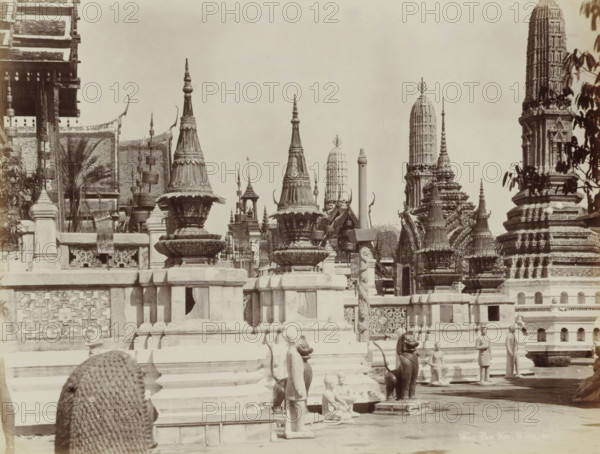 Wat Pra Keo, B.kok, Temple complex of Wat Pra Keo (Temple of the Emerald Buddha) in Bangkok, album page, photograph, Part of Album with 50 photos of a trip through Southeast Asia., check stamp: 'GR Lambert Singapore', photographer: GR Lambert & Co., Bangkok, c. 1870 - c. 1900, paper, albumen print, height 209 mm x width 273 mm, temple, shrine, Hinduism, Buddhism, Jainism, Stone terraces and carved balustrades fill the foreground, topped by ornate bell-shaped and spire-like structures with layered decorative bands and pointed finials. A series of tall, cylindrical towers and sculpted prangs rise in the midground, each covered in intricate relief and patterned surfaces. Several statues and sculpted figures stand or kneel along the steps and platforms, including human forms and animal-like guardians with curved tails and seated postures. Ornamental rooflines with upturned eaves and hanging decorative elements are visible at the left, while leafy branches frame part of the scene at the rig...