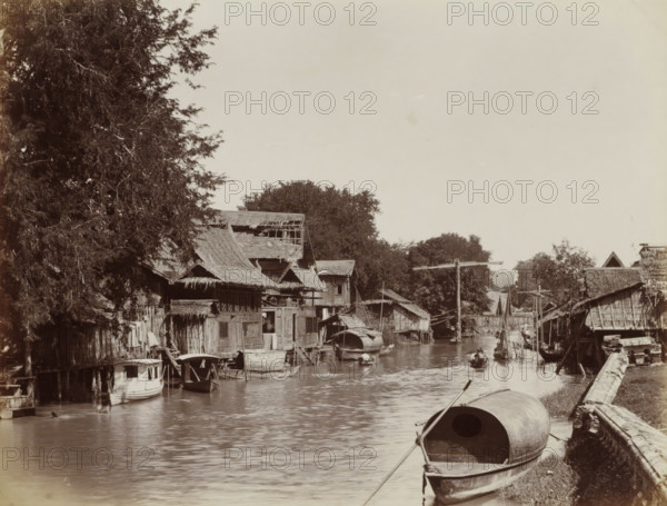Village by a water in Siam, album page, photograph, Part of Album with 50 photos of a trip through Southeast Asia., check stamp: 'GR Lambert Singapore', inscription: 'Siamese Village at Klang San', photographer: GR Lambert & Co., Thailand, c. 1870 - c. 1900, paper, albumen print, height 210 mm x width 274 mm, river bank, A riverside scene with wooden stilt houses and thatched roofs lining both sides of a narrow waterway, small boats and covered canoes moored along the banks, a larger covered boat in the foreground tied to the shore, narrow wooden walkways and ladders connecting houses to the water, leafy trees rising behind the houses, a few wooden poles and simple bridges spanning the canal, reflections of the buildings and boats visible on the water surface, and an open sky occupying the upper part of the image.
