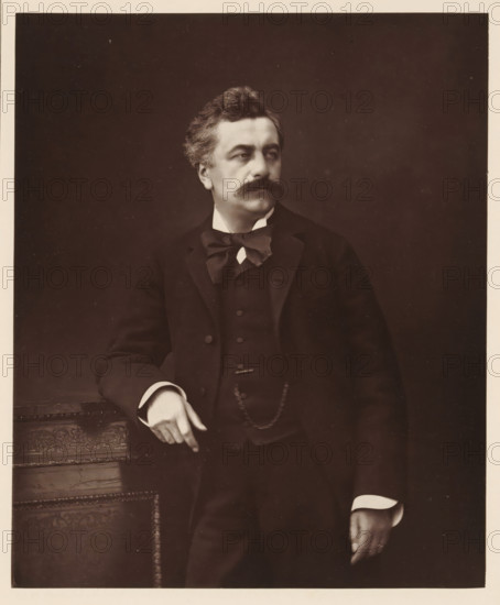 Portrait of Louis Andrieux, L. Andrieux. Ne a Trevoux, le 23 fevrier 1840, page, photomechanical print, inscription: '126, boul. Magenta.-Paris.', photographer: Ferdinand Mulnier, cliché maker: Goupil & Cie., c. 1876 - in or before 1879, paper, Woodburytype, height 225 mm x width 185 mm, Person, Louis Andrieux, politician, eg party leader, A sepia portrait of a man from the waist up wearing a dark three-piece suit with a bow tied at his collar and a visible chain draped across his vest; he has wavy hair, a thick mustache, and is looking slightly to his right; his left arm rests on an ornately carved wooden surface while his right hand hangs by his side showing a ring on a finger; the background is plain and dark.