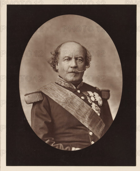 Portrait of François Certain Canrobert, Canrobert. Ne a Saint-Cere (Lot), in June 1809, page, photomechanical print, inscription: '126, boul. Magenta.-Paris.', photographer: Augustin Aimé Joseph Lejeune, cliché maker: Goupil & Cie., c. 1876 - in or before 1879, paper, Woodburytype, Woodburytypepie, height 226 mm x width 181 mm, Person, François Certain de Canrobert, commander-in-chief, general, marshal, A sepia-toned oval portrait of a man shown from the torso up, set against a darker rectangular background. He has a balding head with curly hair at the sides and a prominent curled mustache. He wears a dark military-style uniform with ornate embroidered collar and fringed shoulder epaulettes, a broad sash crossing his chest, and a cluster of decorative medals and badges on his left chest. His expression is neutral and he faces forward.