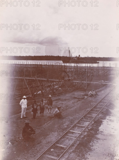 Overview of part of the construction pit and 9th caisson, photograph, Construction site with supervisor and workers, a track on the right. Numbered: 14. Part of the group of loose photographs regarding the construction of the port of Belawan on the coast off Medan, Sumatra 1921-1922., inscription: 'No. 14. Overview of part of the construction pit and 9th caisson', date: '2, 4-21', Belawan, 1921-04-02, photographer, Sumatra, paper, daylight gelatin silver print, height 220 mm x width 160 mm, harbour, building-site, railway, tramway, rack railway, economic history, overseas history, traffic and transport, infrastructure, harbor, Period, 1921 - 1922, A riverside scene with wooden scaffolding and walkways extending along the water, a large boat moored near the far bank, a stretch of rails running along the muddy foreground, several men standing and sitting on the ground wearing hats and light clothing, a pile of goods or materials near the shore, and a distant treeline under a cloudy sky.