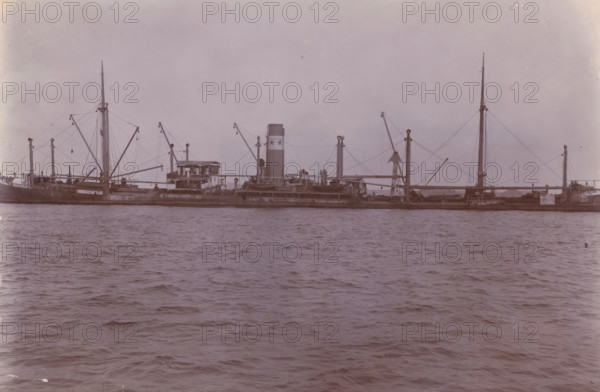 Image of SS Altenburg in Singapore, Belawan Ocean Harbor, photograph, The ship SS Altenburg in Singapore. Numbered: 71. Part of the group of individual photographs regarding the construction of the port of Belawan on the coast off Medan, Sumatra 1921-1922., inscription: 'No. 71, Image of SS Altenburg in Singapore', date: '23, 9-21', Singapore, 1921-09-23, photographer, paper, daylight gelatin silver print, height 160 mm x width 220 mm, steamsailer, economic history, overseas history, traffic and transport, infrastructure, harbor, Sumatra, Belawan, Period, 1921 - 1922, A long steamship viewed from the water with a tall smokestack near the center, multiple masts and booms connected by rigging, a raised wheelhouse and deck structures amidships, lifeboats and cargo gear visible along the deck, calm rippling water in the foreground, and a pale sky above.