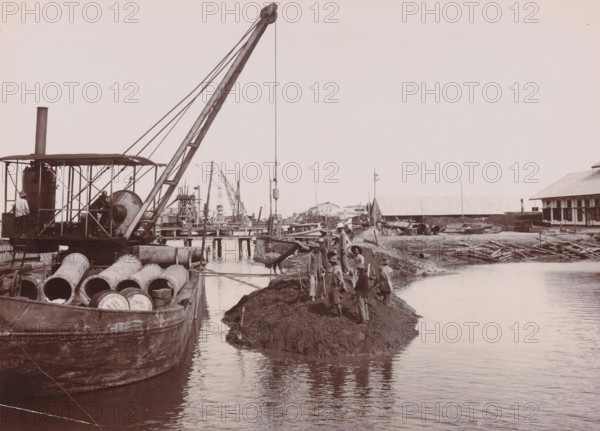 Closing of the dam around the construction pit, Belawan Ocean Harbor, photograph, Closing the dam around the construction pit. Numbered: 102. Part of the group of individual photographs regarding the construction of the port of Belawan on the coast off Medan, Sumatra 1921-1922., inscription: 'No. 102 Closing of the dam around the construction pit', date: '20, 12-21', Belawan, 1921-12-20, photographer, Sumatra, paper, daylight gelatin silver print, height 160 mm x width 220 mm, harbor, building-site, economic history, overseas history, traffic and transport, infrastructure, dredging, Period, 1921 - 1922, A riverside worksite shows a large crane mounted on a barge with a tall boom and cables, a winch and a covered engine area, and several large cylindrical pipes lying on the deck of the barge; a suspended bucket hangs over the water while workers wearing hats and light clothing stand and work on a muddy embankment beside the water, some holding shovels; in the background are wooden piers...