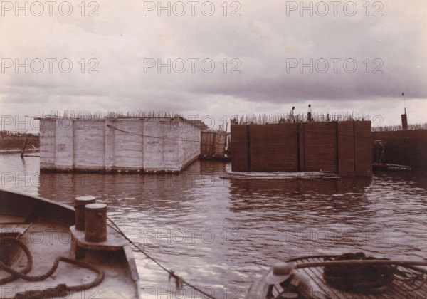 Dismantling, plastering and tarring of the 9.09 m high caissons. (1st work), Ocean Harbor Belawan, photograph, Finishing of the caissons. Numbered: 129. Part of the group of loose photographs about the construction of the harbor Belawan on the coast off Medan, Sumatra 1921-1922., date: '10, 3-22', inscription: '129 Dismantling, plastering and tarring of the 9.09 m high caissons. (1st work)', Belawan, 1922-03-10, photographer, Sumatra, paper, daylight gelatin silver print, height 160 mm x width 220 mm, harbor, building-site, economic history, overseas history, traffic and transport, infrastructure, dredging, Period, 1921 - 1922, A waterscape with large rectangular floating structures framed by wooden and concrete surfaces, vertical metal rods protruding from their tops, two people standing on one of the structures, calm rippling water reflecting the sky, a foreground view of a vessel deck with mooring posts, ropes, and a coil, a small boat tied alongside one of the floating platforms, d...