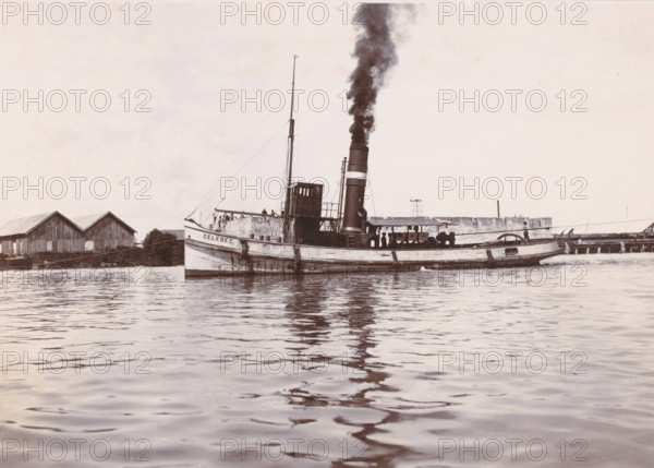 Tugboat 'Celebes', Ocean Harbor Belawan, photograph, The tugboat 'Celebes' with smoking chimney. Numbered: 163. Part of the group of individual photographs regarding the construction of the harbor of Belawan on the coast off Medan, Sumatra 1921-1922., date: '25, 5-22', inscription: 'No 163 Tugboat 'Celebes'', Belawan, 1922-05-25, photographer, Sumatra, paper, daylight gelatin silver print, height 160 mm x width 220 mm, harbor, building-site, tug, pusher tug, economic history, overseas history, traffic and transport, infrastructure, dredging, Period, 1921 - 1922, A steamship with a tall smokestack emitting dark smoke is floating on calm water, its hull showing signs of wear and weathering and a small raised wheelhouse amidships, with several people visible on the deck near railings; a mast with rigging rises above the deck toward the bow, and the stern has a rounded cabin area. Along the shoreline behind the ship there are wooden buildings with pitched roofs, a low pier structure, and s...