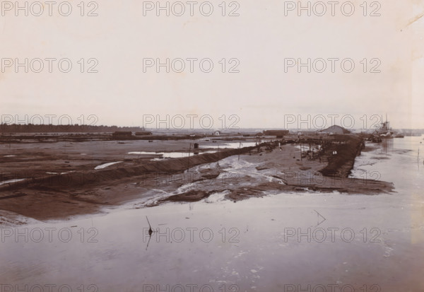 Overview of sand loading, Belawan Ocean Port, photograph, View of the reclaimed land. Numbered: 180. Part of the group of individual photographs regarding the construction of the port of Belawan on the coast off Medan, Sumatra 1921-1922., annotation: 'No. 180 Overview of sand loading', date: '14, 7-22', Belawan, 1922-07-14, photographer, Sumatra, paper, daylight gelatin silver print, height 160 mm x width 220 mm, harbour, building-site, economic history, overseas history, traffic and transport, infrastructure, harbor, dredging, Period, 1921 - 1922, A wide, muddy riverscape fills the scene with shallow pools and meandering wet channels in the foreground, a raised muddy embankment running diagonally toward the right where a cluster of small wooden structures and groups of people are gathered along the water's edge, nearby boats and a larger steamship moored at the far right with visible masts and smokestack, low buildings and warehouses stretch along the midground behind the embankment, ...
