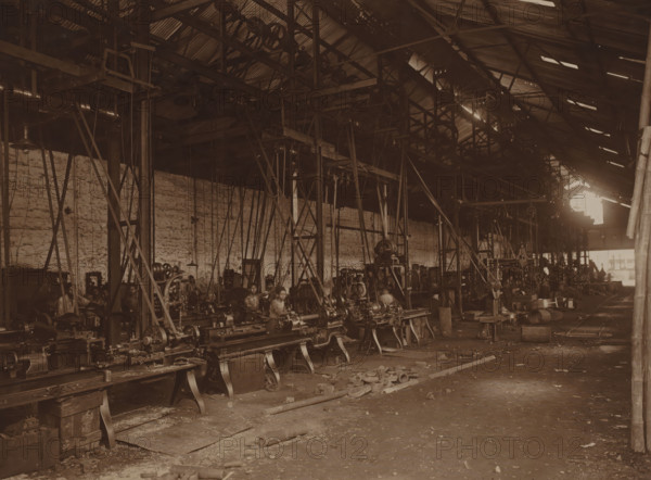 First turning shop, photograph, View into the factory hall with the first turning shop installed in 1904. Pictured here after the electrification in 1908. Part of the binder with photos of machines from NV Machinefabriek Braat in Surabaya, Sukabumi, and Yogyakarta, from the period 1908-1942., Surabaya, 1908, photographer, Java, East Java, paper, photography, height 153 mm x width 202 mm, Person, NV Machinefabriek Braat, colonial system, factory-interior, machine shop, machines, parts of machines, tools and appliances, economic history, overseas history, business history, Period, 1908 - 1942, A large industrial interior with rows of heavy metal lathes and machinery arranged on raised supports, a high corrugated metal roof with exposed beams and pulley systems, vertical and diagonal metal supports and belts hanging down toward the machines, a long brick wall along one side, several workers seated or standing at the machines, scattered metal parts and debris on the concrete floor, shafts ...