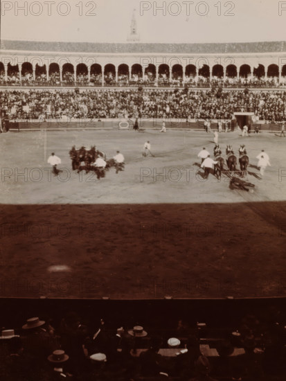 At the Bull Fight, Spain, photograph, Arena for bullfights in Valencia or Barcelona., 1905 - 1922, photographer, paper, gelatin silver print, developing gelatin silver print, height 105 mm x width 85 mm, Period, A large oval arena with a dirt floor fills the center of the image, with groups of people and teams of horses on the arena surface; several figures in light clothing are near the horses, some standing and some appearing to move, and there is a fallen figure or object near one team of horses. Surrounding the arena is a multi-tiered colonnade with arches filled with a dense crowd of seated spectators, and a tall central tower rising above the roofline in the background. In the foreground the lower seating area is visible in shadow, showing spectators wearing hats and seated closely together, with portions of wooden benches and the top of a protective barrier separating the seating from the arena. The overall image has a sepia tone and shows signs of age with slight fading and gra...