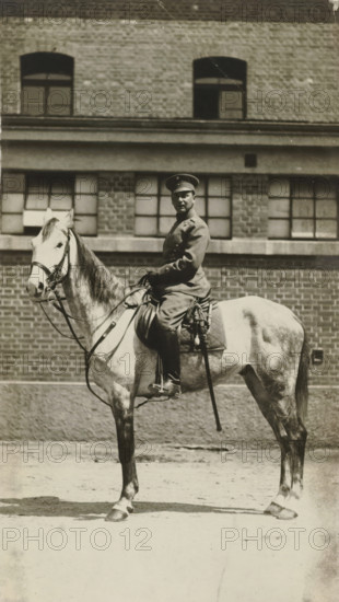 Arthur Moos on horseback in the uniform of the 20th Uhlanen regiment, May 1916, in Ludwigsburg, photograph, Arthur Moos on horseback in the uniform of the 20th Uhlanen regiment in Ludwigsburg., Ludwigsburg, 1916-05, photographer, Baden-Württemberg, paper, gelatin silver print, developing gelatin silver print, height 135 mm x width 80 mm, Person, Arthur Moos, Period, A black-and-white photograph showing a person in a uniform and cap seated on a light-colored horse in front of a brick building with multi-pane windows; the rider faces slightly toward the camera and holds the reins, wearing boots and a belted coat with visible buttons, and a sword or saber hangs at the saddle; the horse has a bridle and saddle with straps and stands on a paved surface, while the building behind shows rows of bricks, window frames, and a horizontal ledge.