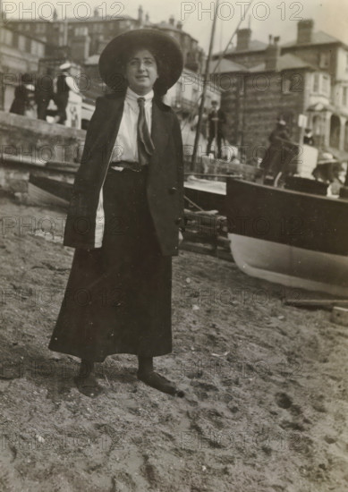 Else Wachenheimer-Moos with a hat in front of a boat on the beach, Bexhill-on-Sea, 1913, photograph, Bexhill-on-Sea., photographer, East Sussex, paper, gelatin silver print, developed gelatin silver print, height 110 mm x width 80 mm, Person, Else Wachenheimer-Moos, England, Period, A person stands on a sandy shore wearing a long dark skirt, dark jacket, light blouse with a scarf tied at the neck, and a wide-brimmed hat, with dark shoes visible on the sand; behind them a boat is beached with its hull partly visible and ropes and masts nearby, and further back there are buildings on a raised quay with several other figures and architectural details along the waterfront.