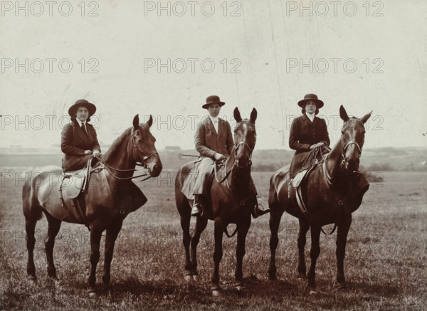 Else Wachenheimer-Moos and two unknown women on horseback, Bexhill-on-Sea in England, 1913, photograph, Else Wachenheimer-Moos (left) and two unknown women on horseback, Bexhill-on-Sea in England., Bexhill-on-Sea, photographer, East Sussex, paper, gelatin silver print, developing gelatin silver print, height 240 mm x width 310 mm, Person, Else Wachenheimer-Moos, England, Period, Three people wearing dark riding coats and wide-brimmed hats sit astride three dark horses in a grassy field; the riders hold reins and one holds a crop, the horses wear saddles and bridles and face forward with visible ears and manes, and the background shows a pale sky above a distant, low horizon.