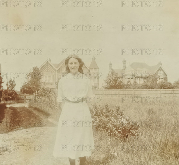 Marjorie in a rural setting with a building in the background in Bexhill-on-Sea in England, 1913, photograph, Marjorie in a rural setting with a building in the background in Bexhill-on-Sea in England., inscription: 'Marjorie', Bexhill-on-Sea, photographer, East Sussex, paper, gelatin silver print, developed gelatin silver print, height 90 mm x width 80 mm, England, Period, A young woman stands outdoors in a light-colored dress with sleeves rolled to the elbows and a belt at her waist, her hair parted and loose around her shoulders and she faces the camera with a neutral expression; behind her is a wooden fence and beyond that a row of large houses with steep gables, chimneys, and turrets set among trees and shrubbery, and in the foreground there is a dirt path and areas of grass and low plants.