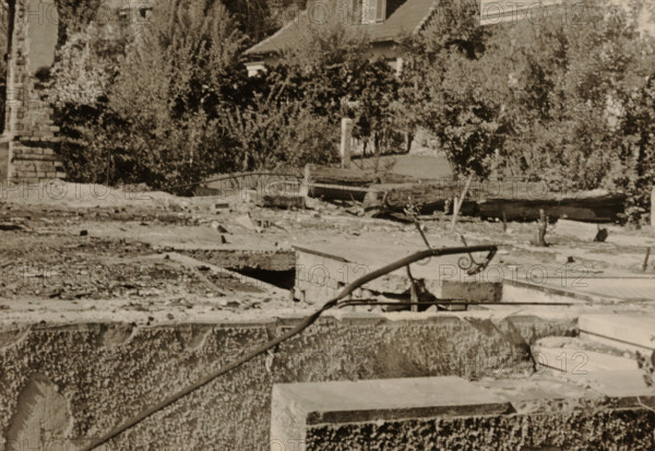Top of part of a ruin, with a tree-lined house in the background, photograph, with a tree-lined house in the background., 1945 - 1970, photographer, paper, gelatin silver print, developed gelatin silver print, height 75 mm x width 105 mm, Period, A sepia-toned scene showing a rough concrete foreground with exposed edges and a curved metal handrail topped with a decorative spiral, rubble and broken slabs scattered across a flat midground with openings and gaps, a low stone wall and a rusted pipe lying across the surface, and dense shrubs and trees behind with partial views of a tiled roof and a window peeking through the foliage.