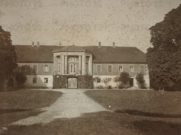 Front view of a country house with a gazebo-shaped central section, with trees on either side and a lawn and driveway at the front, photograph, with trees on either side and a lawn and driveway at the front., 1900 - 1950, photographer, paper, gelatin silver print, developed gelatin silver print, height 80 mm x width 105 mm, Period, A sepia-toned photograph of a large two-story building with a central portico supported by columns and an arched passage at ground level, flanked by windows on both sides and a pitched roof with chimneys; a gravel path leads from the foreground toward the archway, with lawns on either side and tall trees partially framing the building at the left and right edges of the image.
